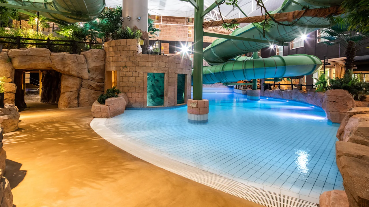En stor grøn vandrutsjebane bøjer sig over en klarblå pool i en indendørs vandpark. Området er omgivet af kunstige klippeformationer og planter.