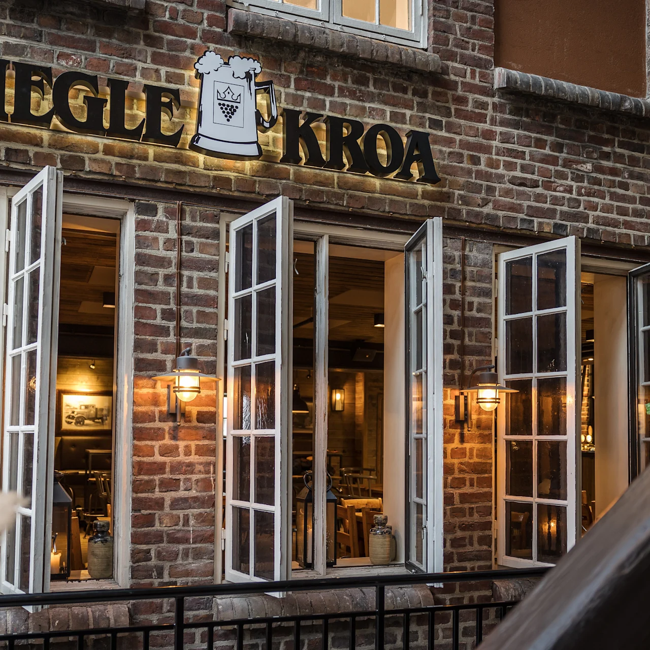 Tiilitalo, jossa on avoimet ikkunat ja viihtyisä sisustus. Kyltissä lukee "KIEGLE KROA" olutmukin kera, ympärillä vehreyttä ja lämmin valaistus.