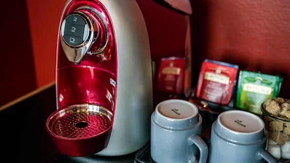 A red and silver coffee machine is on a black counter, surrounded by assorted tea bags, two gray mugs, and a small bowl of sugar cubes. 