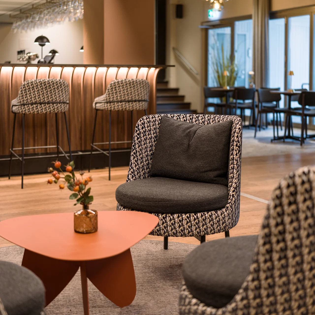 Comfortable chairs encircle a small, modern table with a plant, in a stylish lounge with a bar and high stools in the background.
