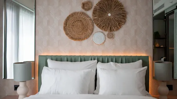 A neatly made bed with white pillows and a green headboard, flanked by lamps, under a woven circle wall art. Soft light glows behind the headboard. 