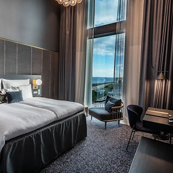 En stor seng med hvidt sengetøj er placeret ved siden af et gulv-til-loft vindue med udsigt over havet. Rummet omfatter et skrivebord, stol og moderne belysning.