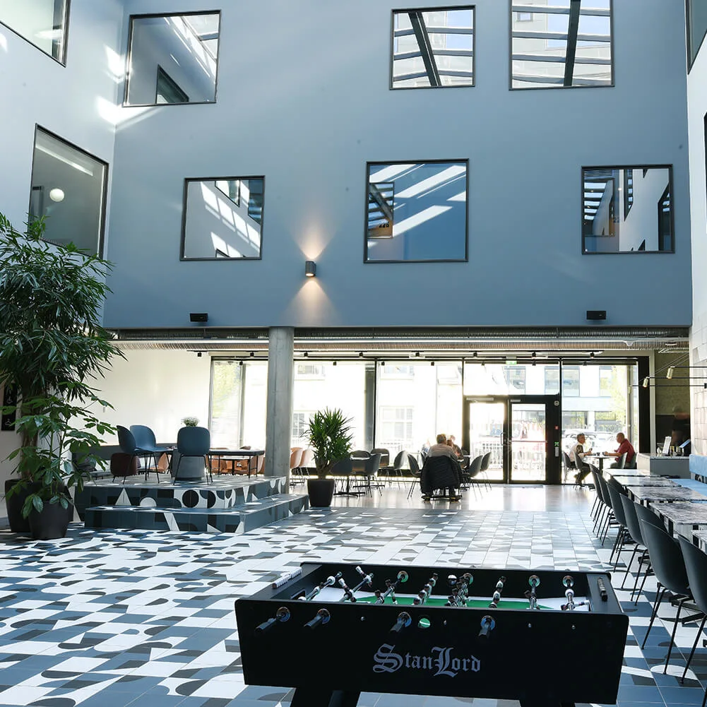 Bordfodboldbord i en rummelig, moderne lobby med flisemønstret gulv, blå vægge og store vinduer. Folk sidder ved borde tæt på receptionen. Tekst på bordfodbold: "Stanlord."