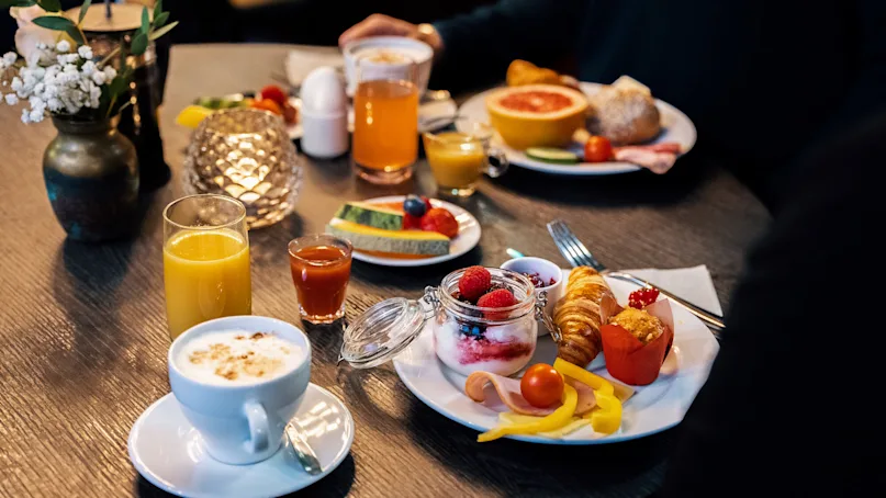 Breakfast spread featuring a cappuccino, orange juice, fruit, yogurt parfait, croissant, cold cuts, and vegetables on a dark wooden dining table.