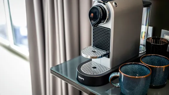 En kompakt silverfärgad kaffemaskin står på ett glasbord, omgiven av två blå keramikkoppar. Text: "KRUPS NESPRESSO" syns på maskinens bas.