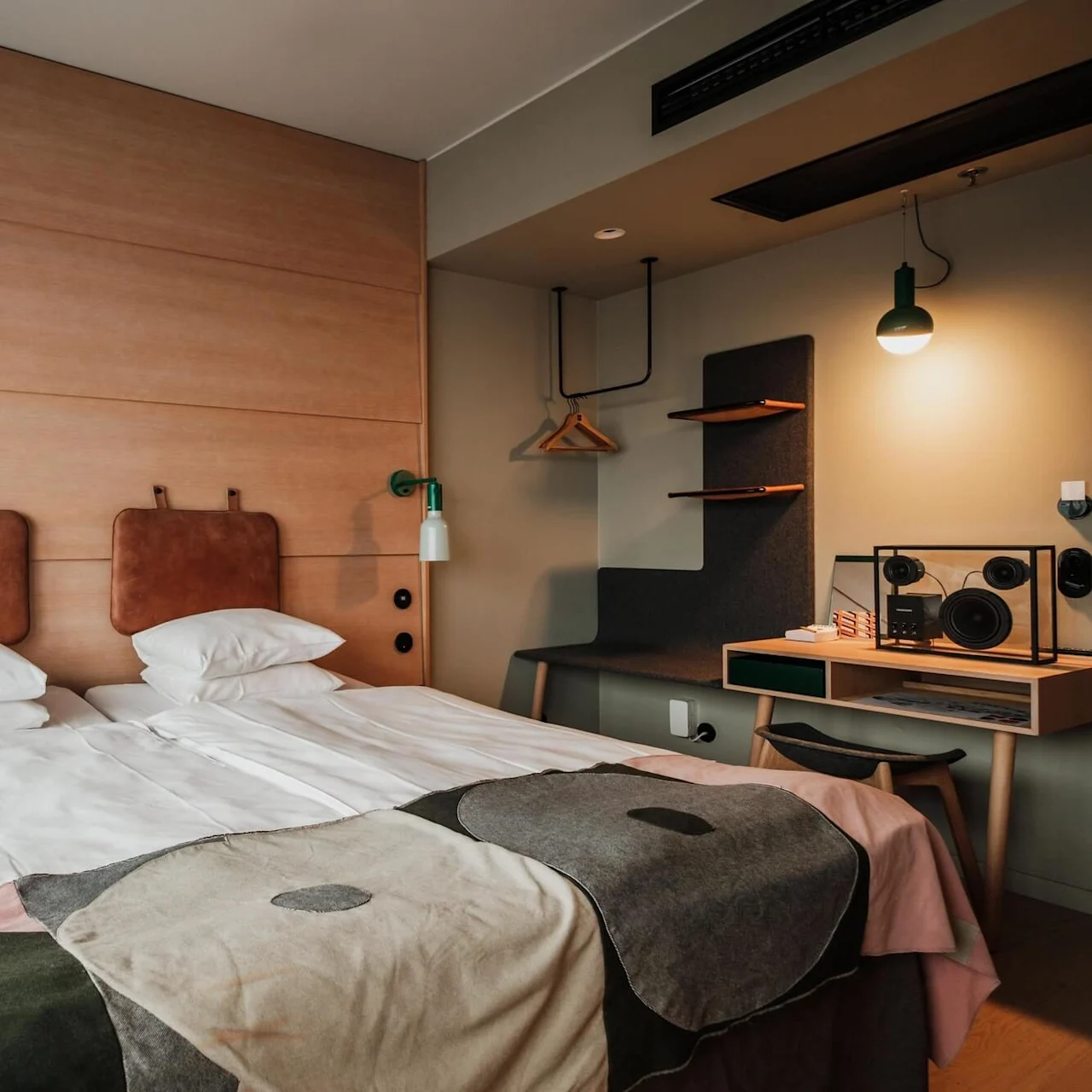 A neatly made double bed with white pillows and patterned blanket is in a modern hotel room. Wooden side table, red lamp, and desk area with lighting are nearby.