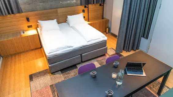 A neatly made double bed with white linens in a modern room. A table with a laptop, cups, and a water bottle sits in the foreground. Windows provide natural light.