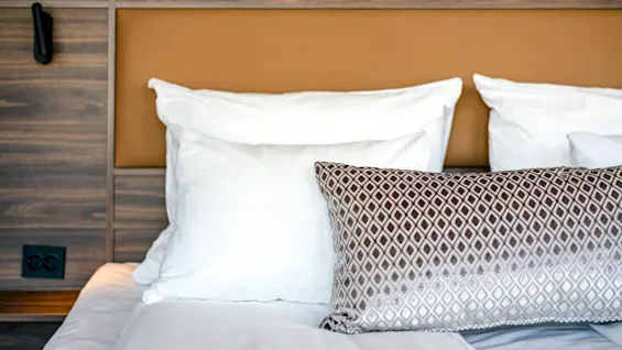 A bed with white pillows and a patterned bolster rests against a wooden headboard. A black wall lamp and electrical outlet are nearby, suggesting a hotel room setting.