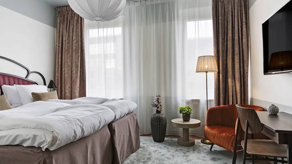 Lyst og koselig hotellrom med oppredd dobbeltseng, sittegruppe ved vinduet og et lite skrivebord.
