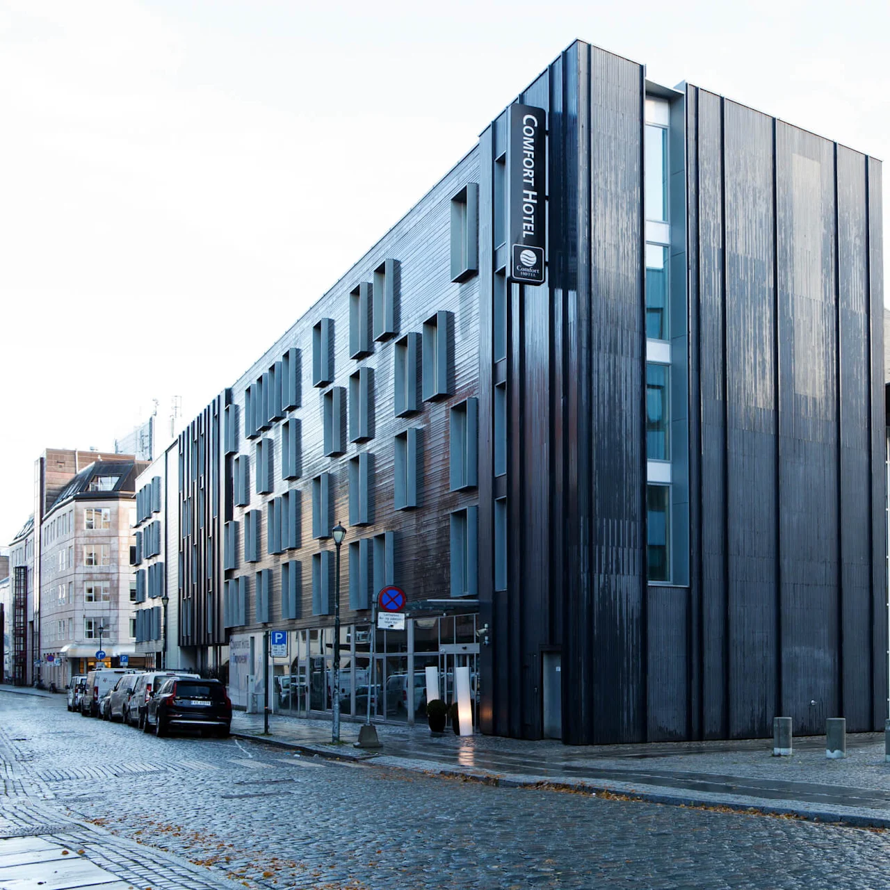 En modern byggnad med en mörk fasad, som visar skylten "COMFORT HOTEL" vertikalt. Den ligger längs en gatubeläggning med parkerade bilar och angränsande byggnader.