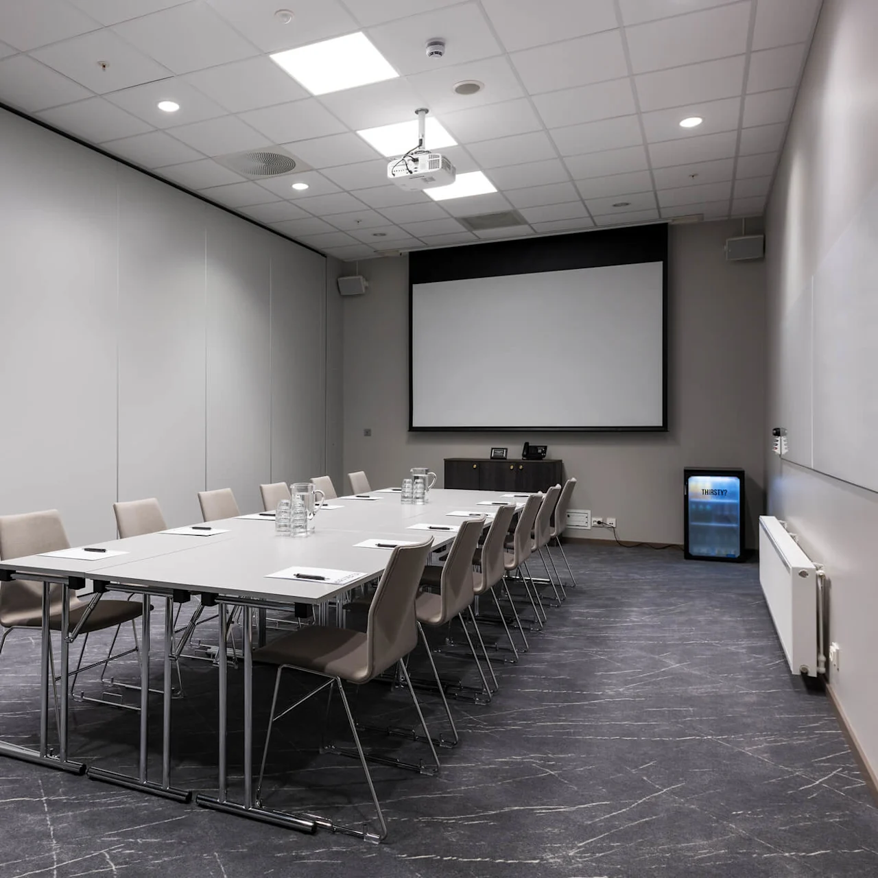 Et langt konferansebord med stoler er satt opp med papirer og vannkarafler i et moderne møterom, med en stor skjerm, projektor og et mini-kjøleskap merket "FROST."