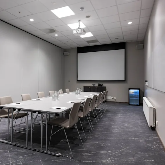 Et langt konferansebord med stoler er satt opp med papirer og vannkarafler i et moderne møterom, med en stor skjerm, projektor og et mini-kjøleskap merket "FROST."