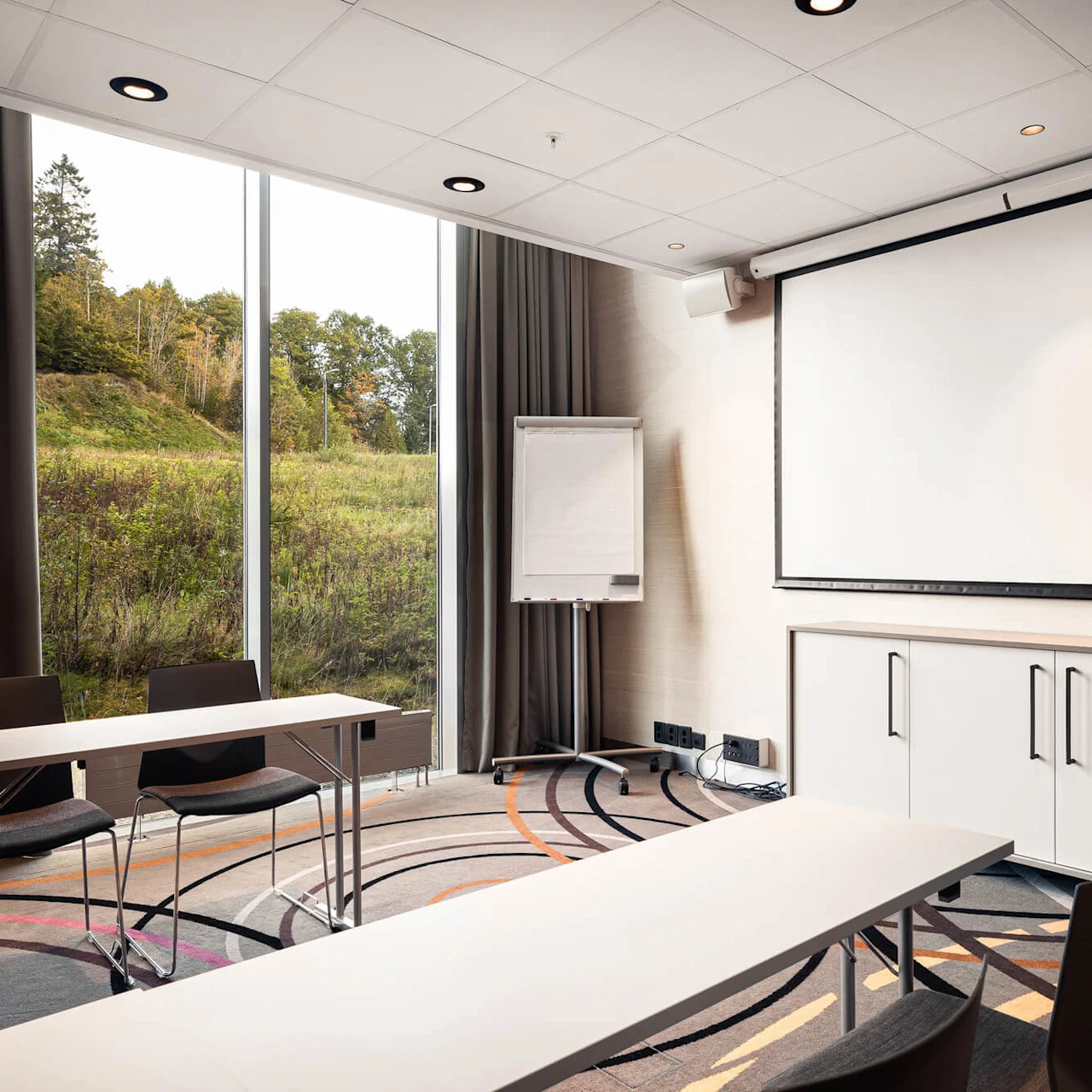 A conference room features a white projection screen, tables, chairs, and windows with a view of trees and grass outside. A flip chart stands in the corner.