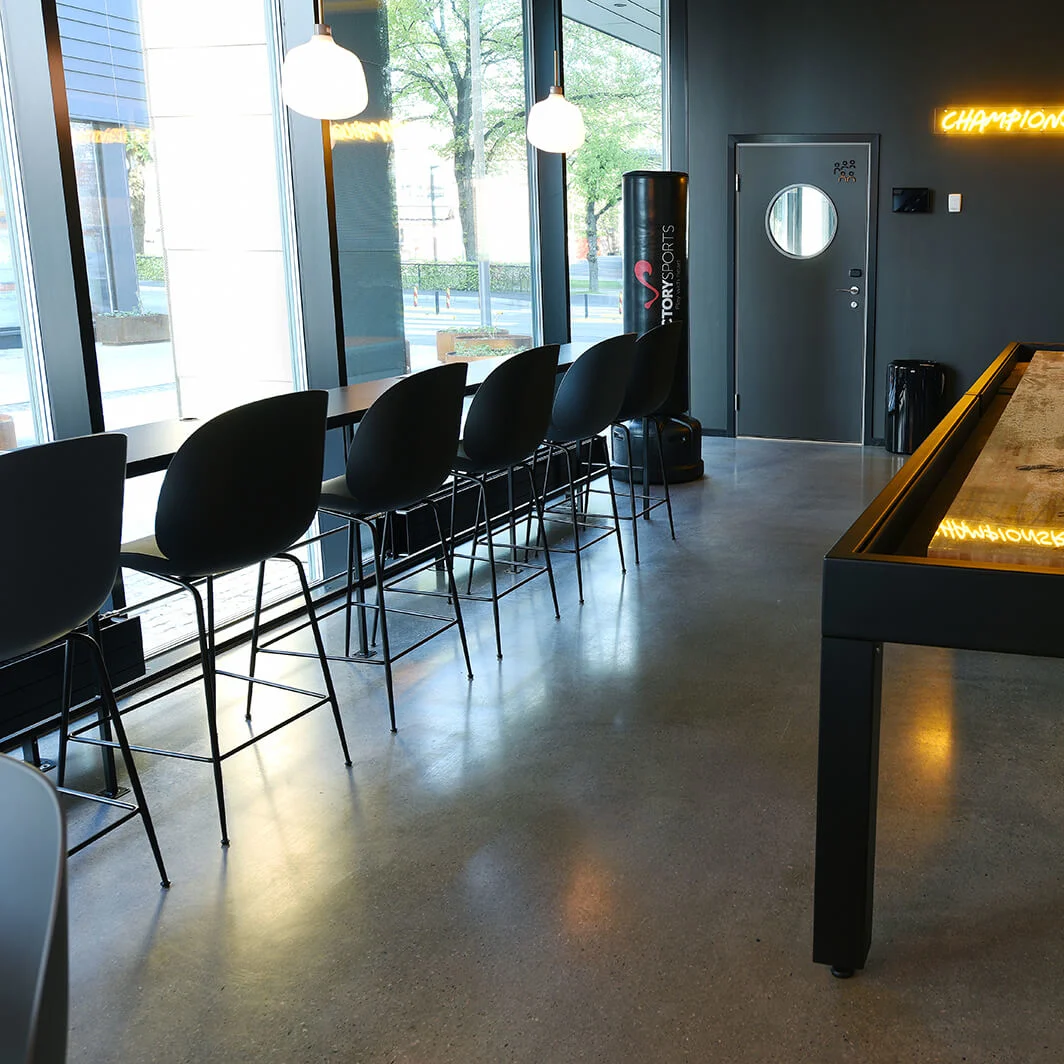 Shuffleboard på et sort bord i en moderne lounge med høje stole ved store vinduer. Neonskilt med teksten "CHAMPIONSROOM."