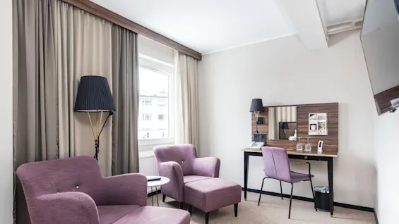 Två lila fåtöljer med fotpall och golvlampa står i ett hotellrum med skrivbord, gardiner och fönster.