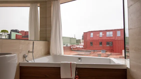 Ett modernt badrum med ett badkar vid ett stort fönster med utsikt över stadens takåsar. Vita gardiner ramar in utsikten.