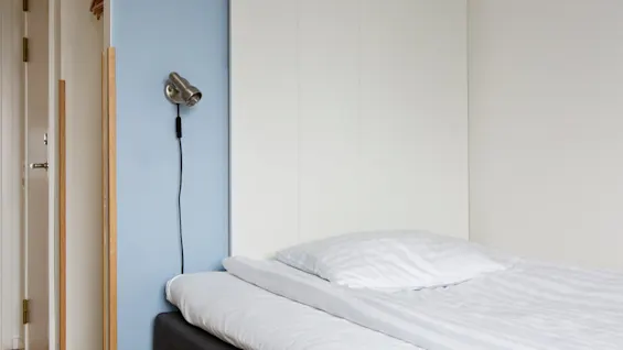 A neatly made bed with white linens sits in a minimalist room. A wall-mounted silver lamp with a black cord is on a light blue wall next to the bed.