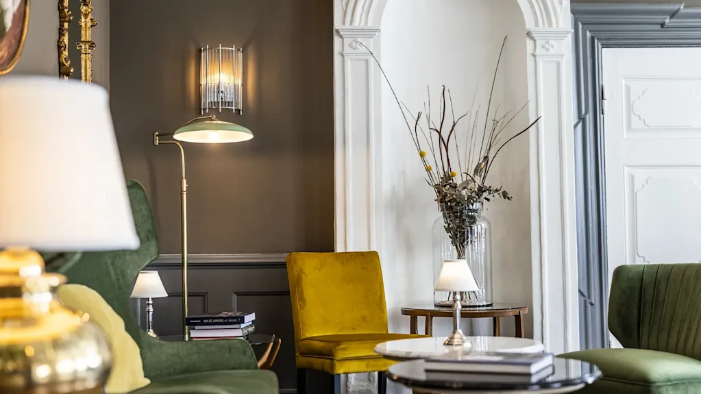 En hyggelig hotellounge med grønne og gule stole, lamper og et sofabord, der skaber en indbydende atmosfære.