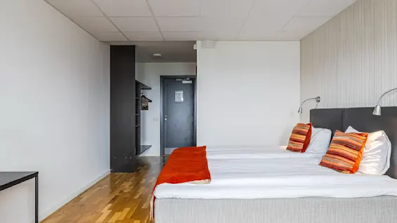 Et hyggelig hotellrom med to enkeltsenger, hvitt sengetøy og fargerike puter. Et tregulv og enkel innredning gir varme. En åpen garderobe og en dør er synlig i bakgrunnen.