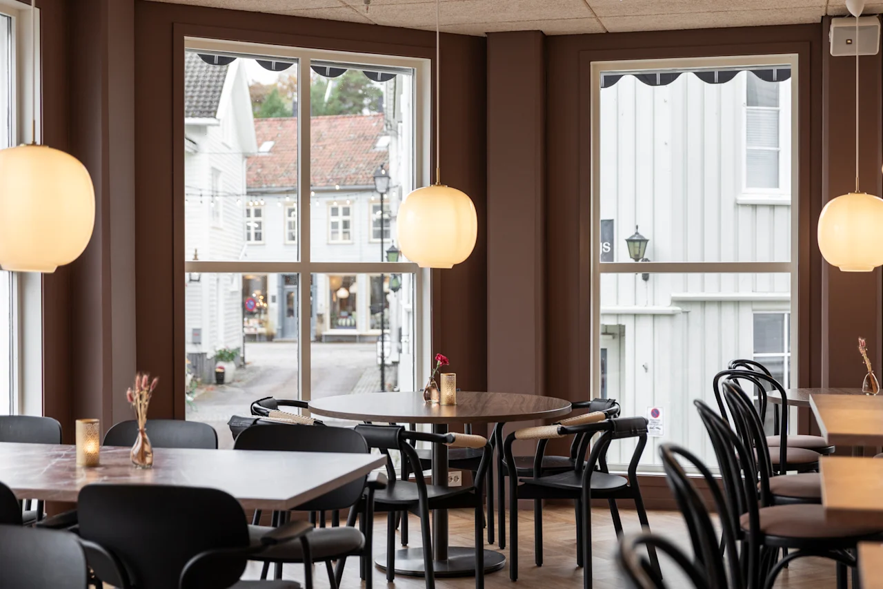 A warm and inviting cafe interior with large windows offering a view of a quaint street, furnished with tables and chairs.