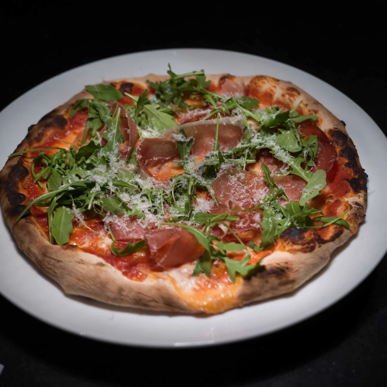 A pizza with prosciutto, arugula, and grated cheese rests on a white plate. It is set against a dark surface, illuminated by a spotlight.