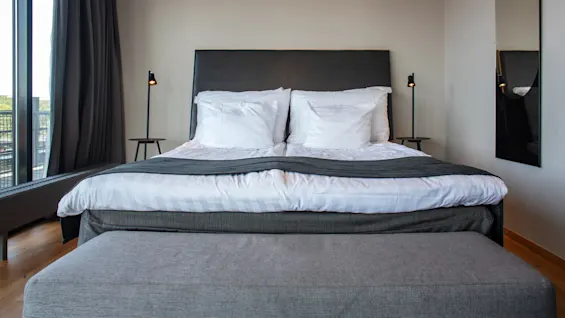 A neatly made double bed with white pillows and bedding is flanked by matching bedside lamps. It faces a tall window with dark curtains in a modern bedroom.