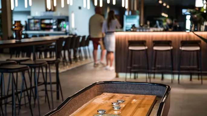 Shuffleboard, jossa on metallisia pelimerkkejä, lepää puupinnalla, modernissa baarimiljöössä, jota ympäröivät korkeat tuolit ja keskusteleva ihmisjoukko taustalla.