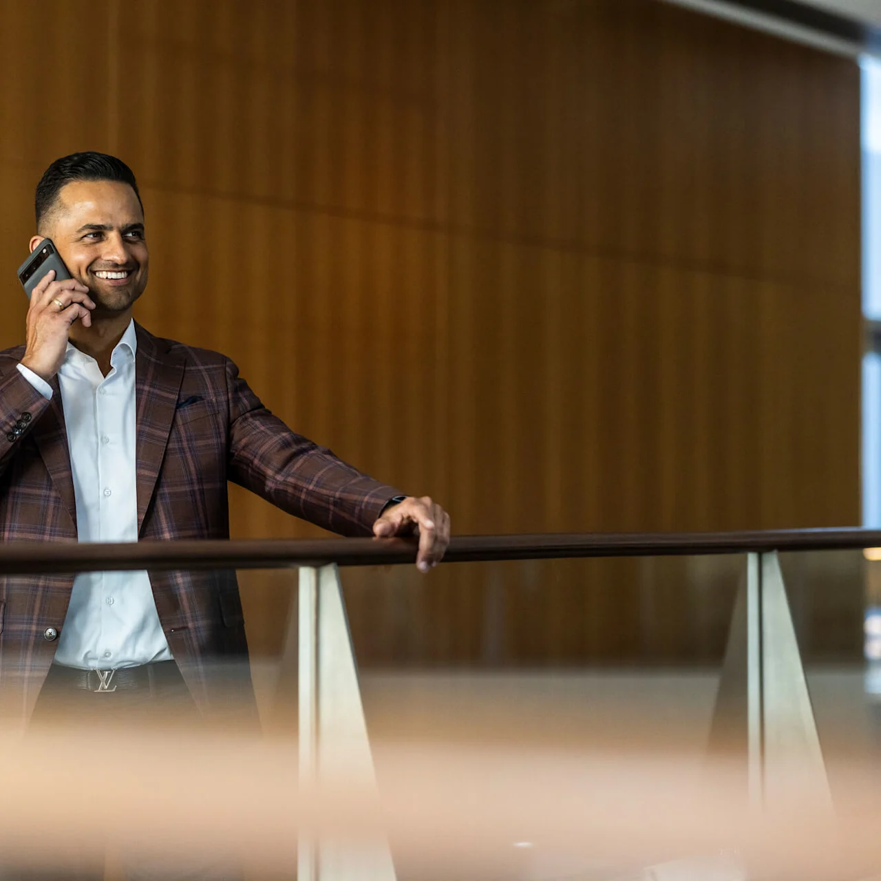 En man i en rutig blazer pratar i mobiltelefonen och ler, stående vid ett räcke i en modern, träpanelad interiör med stora fönster.