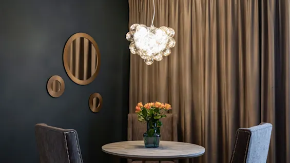 A cozy dining area at Home Hotel Grand Bodø with a round table, chairs, a vase of flowers, and a unique light fixture.