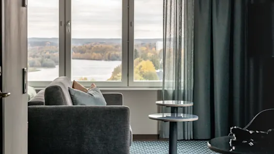 Grå sofa vender mod stort vindue med udsigt over efterårstræer ved en sø; runde borde ved siden af sofa; rum med blå gardiner og mønstret tæppe.