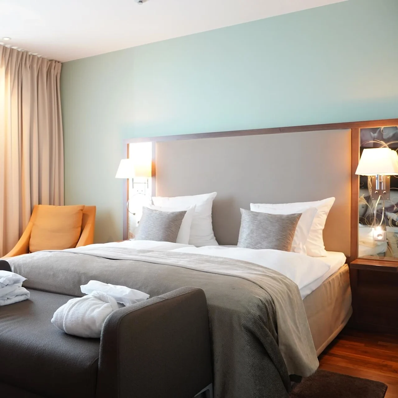 A bed with white and gray pillows stands neatly arranged. Nearby, an orange chair and round table with flowers complete the serene, softly lit room.