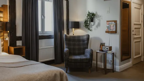 Et hyggeligt hotelværelse med en ternet lænestol, en seng og et vindue. Et magasinholder på væggen viser 'Evigt ung'.