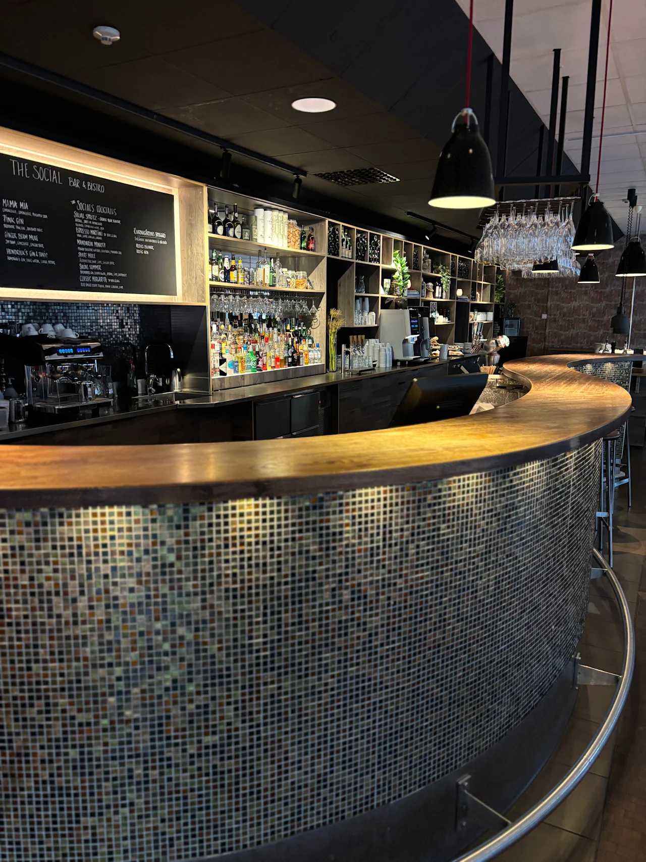 Modern bar med böjd trädisk, kaklad front och flaskhyllor. Menyn: THE SOCIAL BAR & BISTRO SOCIALS COCKTAILS.