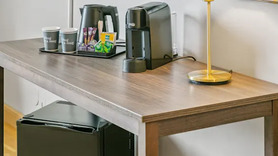 En kaffe station på et træbord har en sort kaffemaskine, en elkedel, to papirkopper mærket "A hug in a mug," og forskellige kaffeartikler, ved siden af et mini-køleskab og lampe.