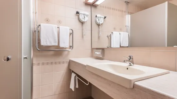 A beige tiled bathroom features a white sink, two wall-mounted hairdryers, and towel racks with neatly hung towels, offering a clean and organized appearance.