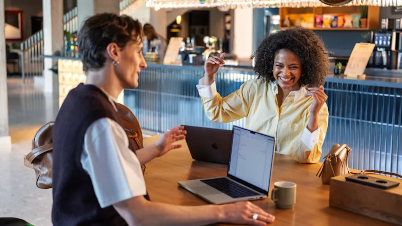 En kvinna och en man samtalar livligt vid ett träbord med bärbara datorer i ett modernt, varmt belyst kafé. Glas och snacks pryder baren i bakgrunden.