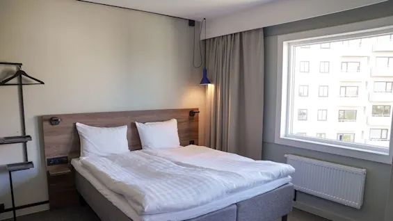 En pent og oppredd dobbeltseng står i et moderne rom, med en trehodegavl og to puter, opplyst av en blå pendellampe, ved siden av et stort vindu med gardiner.
