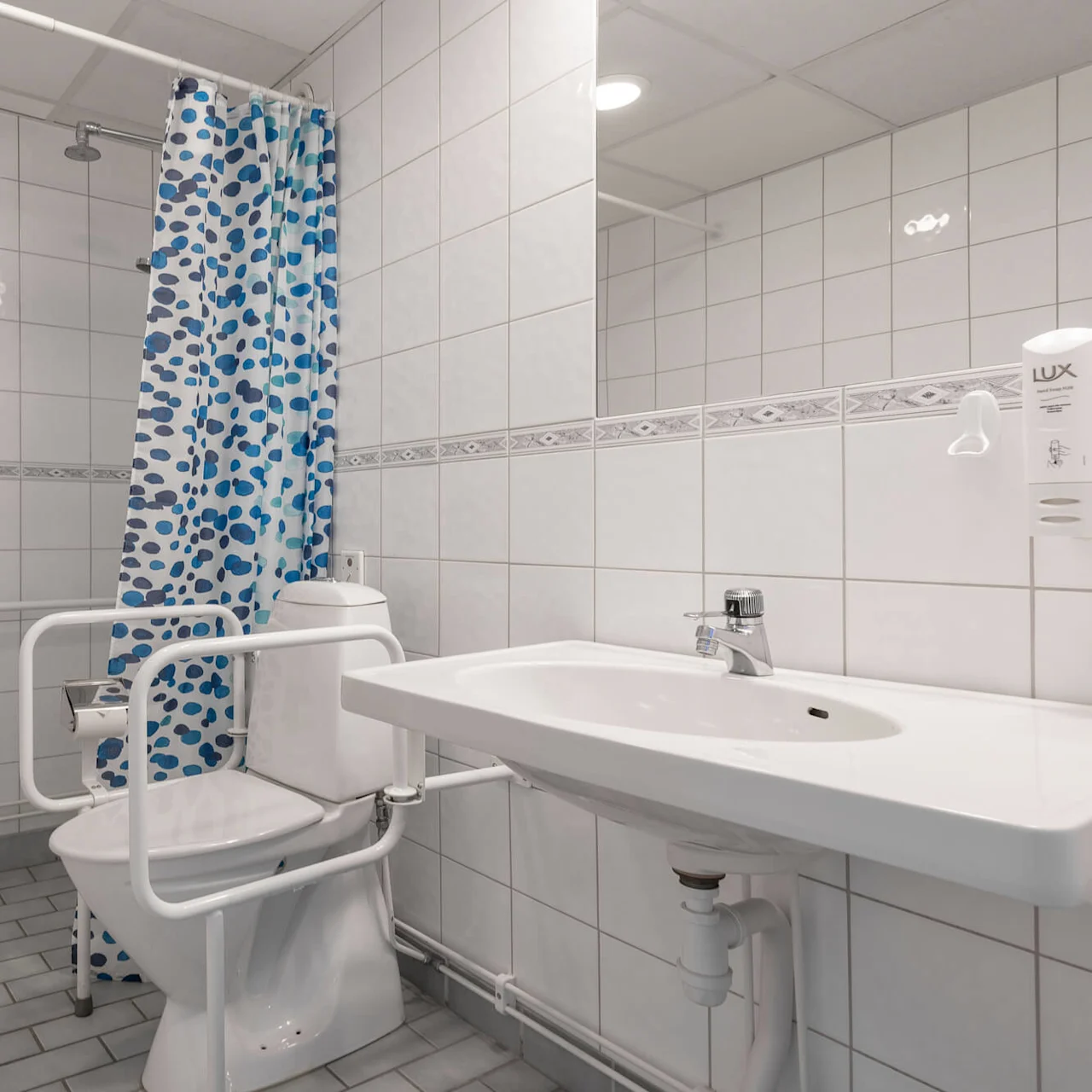 Toalett med säkerhetsräcken är placerad bredvid ett handfat; ett blåmönstrat duschdraperi hänger i närheten. Grått golv och vita väggar skapar ett rent, tillgängligt badrum. Text: "LUX".