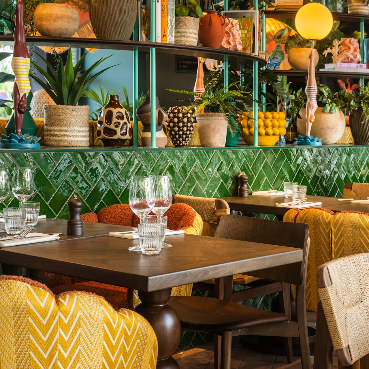 Tables set with glasses and napkins, surrounded by patterned chairs in a colorful restaurant filled with plants and decorative items on green tiled walls.