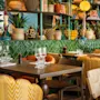 Borde med glas og servietter, omgivet af mønstrede stole i en farverig restaurant fyldt med planter og dekorative genstande på grønne fliser.