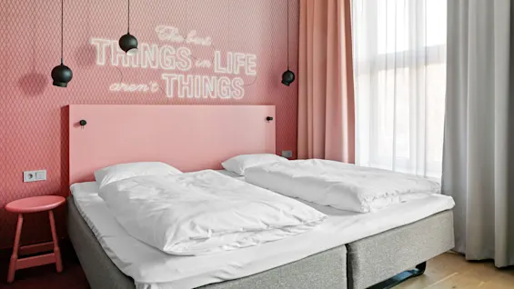 Seng med hvitt sengetøy ved siden av rosa vegg med glødende tekst "The best things in life aren't things," med hengende svarte lamper, ved siden av vindu med rosa og hvite gardiner.
