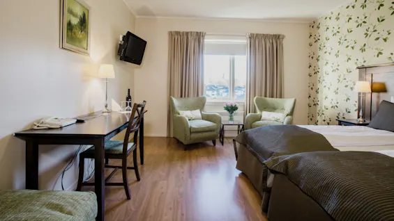 Ett prydligt hotellrum med tvillingbäddar, ett skrivbord med telefon, stolar, blommig tapet, och en väggmonterad TV.
