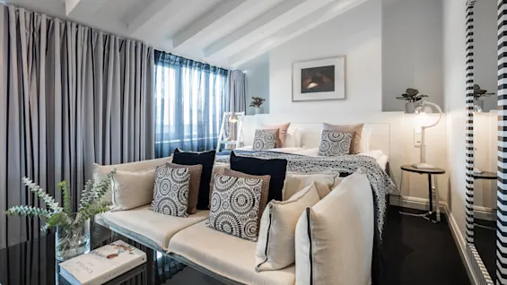 Et moderne soverom har en seng med mønstrede puter, en koselig sofa og store vinduer med gardiner. En speilvegg og et sidebord med lampe tilfører eleganse.