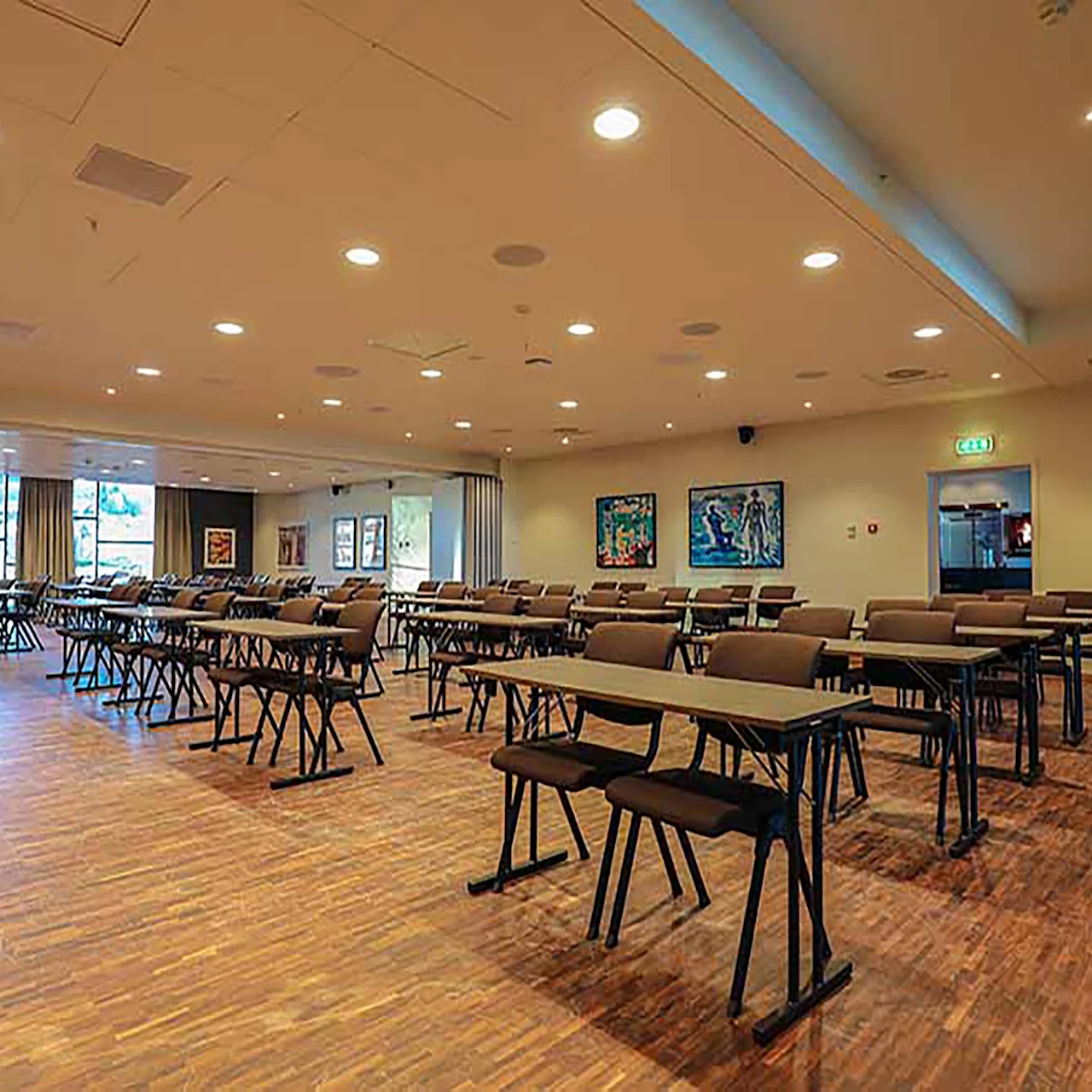 Rader av stoler og bord er ordnet i et romslig, lyst konferanserom med tregulv og moderne malerier på veggene.