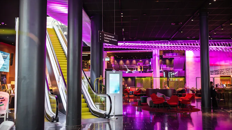 Escalator ascending, surrounded by a vibrant, modern lounge with pink and purple lighting. Seating areas with red chairs and tables. Signage: "Eix ambrosi arribades. Connection flights."