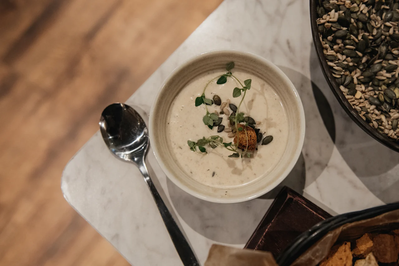 Cremet suppe garneret med urter og frø, med en ske og en skål med frø. Tilføj middag på Home Hotel Norre Park i Halmstad.