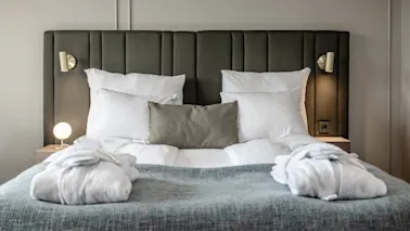 A neatly made bed with white linens and a gray throw, flanked by dimly lit wall lamps in a tranquil bedroom setting.