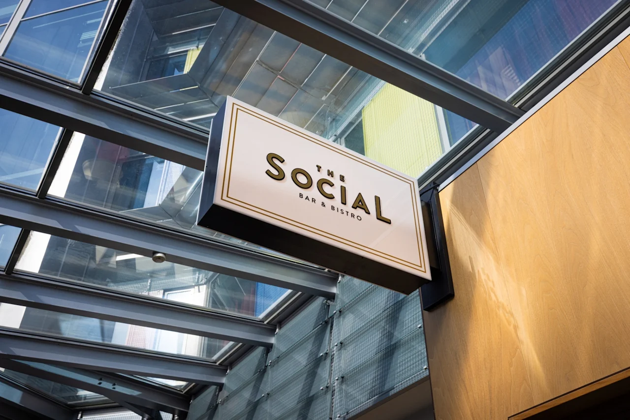Et skilt for "THE SOCIAL BAR & BISTRO" henger under et glasstak, vinklet mot en trevegg i et lyst, moderne miljø.