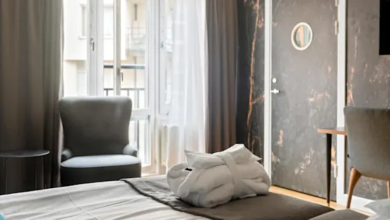 A neatly made bed with a white folded robe on it, in a well-lit hotel room with modern furnishings and balcony doors.