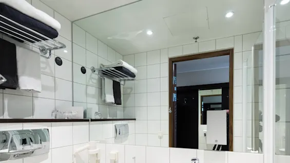 Hvitt keramisk servant er montert på en flislagt vegg på et moderne bad. Det er omgitt av et toalett, speil, håndklekroker og en hårføner, og skaper en ren, lys atmosfære.